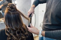 Обрезать неузнаваемый мужчина стилист в повседневной одежде, используя выпрямитель волос, делая кудри для клиента женского пола в салоне красоты — стоковое фото