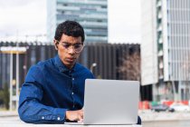 Adulto concentrato etnico maschio dipendente remoto in auricolare che lavora su netbook in città nella giornata di sole — Foto stock