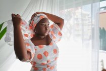 Concentrated young African lady in stylish summer dress adjusting traditional turban while standing near window in light room — Stock Photo