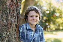 Счастливый стильный маленький мальчик в шортах и клетчатой рубашке опираясь на ствол дерева и улыбаясь во время отдыха на заднем дворе в солнечный день — стоковое фото