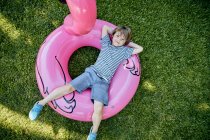 Full body of cheerful little boy in casual clothes lying on inflatable pink flamingo while having fun on grassy lawn in park — Stock Photo