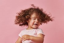 Jolie fille gaie tout-petit avec des cheveux bouclés dans des vêtements décontractés secouant la tête avec les yeux fermés sur fond rose — Photo de stock
