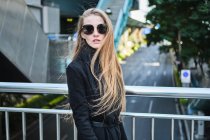 Vista laterale della giovane donna elegante in occhiali da sole e vestito nero sul ponte sulla strada della città — Foto stock