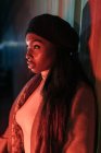 Vista lateral da mulher afro-americana elegante que brilha pela luz vermelha do néon que está perto do edifício na cidade e olhando afastado — Fotografia de Stock