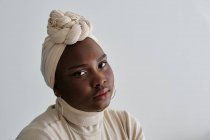 Hermosa joven modelo femenino africano en elegante turbante tradicional de pie sobre fondo blanco y mirando a la cámara - foto de stock