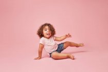 Menina da criança alegre bonito com cabelo encaracolado em roupas casuais se divertindo olhando para longe sorrindo enquanto sentado em fundo rosa — Fotografia de Stock