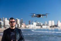 Homem sério em óculos de sol com controlador remoto em pé no drone de voo de praia no litoral perto da cidade — Fotografia de Stock