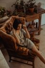 Сверху мирная женщина сидит в потрепанном кожаном кресле и трогает голову, расслабляясь дома — стоковое фото