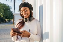 Delighted black female with headphones on neck leaning on building and browsing mobile phone on city street — Stock Photo