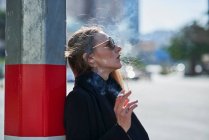 Vue latérale de la jeune femme en écharpe fumant la cigarette près du poteau sur la route urbaine dans rétro-éclairé — Photo de stock