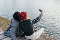 Mulher irreconhecível tomando selfie de amigo feminino preto enquanto relaxa no dique na cidade — Fotografia de Stock