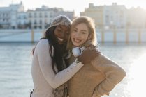 Looking at camera two multiracial female friends in headphones enjoying together on embankment on sunny day — Stock Photo