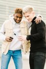 Feliz chico afroamericano joven en ropa de moda sonriendo mientras está de pie en la calle con la mano en el hombro de un amigo varón mostrando video en el teléfono inteligente - foto de stock