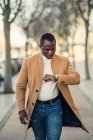 Concentrato giovane afroamericano maschio in abiti alla moda controllare il tempo sul orologio da polso mentre si cammina sulla strada della città nella giornata di sole — Foto stock