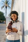 Entzückte schwarze Frau mit Kopfhörern am Hals lehnt an Gebäude und surft auf der Straße — Stockfoto