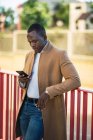 Concentrated young African American male in trendy outfit leaning on fence on street and browsing mobile phone on sunny summer day — Stock Photo