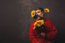 Kreativer reifer Mann im Strickpullover, der das Gesicht mit hellen Sonnenblumen vor schwarzem Hintergrund bedeckt — Stockfoto