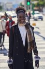 Giovane donna afroamericana di tendenza felice con zaino e occhiali da sole passeggiando sulla carreggiata urbana mentre guarda la fotocamera alla luce del sole — Foto stock