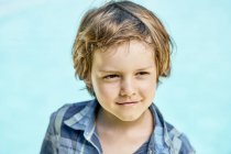 Красивый маленький мальчик с светлыми волосами в стильной клетчатой рубашке улыбается и смотрит в сторону, стоя на синем фоне в солнечном свете — стоковое фото