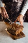 Crop anonymous female baker with knife cutting loaf of fresh bread with sunflower seeds on table in bakehouse — Stock Photo