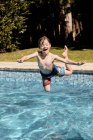 Alegre niño sin camisa gritando mientras salta al agua de la piscina durante las vacaciones de verano en el campo - foto de stock