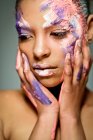 Modelo feminino étnico criativo com rosto manchado com tinta rosa e branca tocando pescoço olhando para baixo no fundo cinza no estúdio — Fotografia de Stock