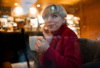 Через стеклянный боковой вид улыбается молодая женщина в уютном красном свитере наслаждаясь ароматным горячим напитком и трогательным лицом, сидя на удобном кресле в кафетерии и мечтательно глядя в сторону — стоковое фото