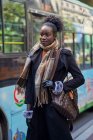 Молода модна етнічна жінка в пальто і шарфі з афро-волосиною булочкою з нетерпінням чекає на міський тротуар — стокове фото
