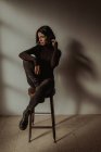 Тонка жінка в чорному вбранні, що сидить на дерев'яному табуреті з піднятими ногами в кімнаті проти білої стіни — стокове фото