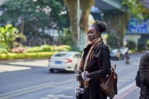 Jeune femme ethnique à la mode en manteau et écharpe avec un chignon afro sur le trottoir urbain — Photo de stock