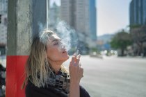 Vista laterale della giovane femmina in sciarpa fumare sigaretta vicino post sulla strada urbana in retroilluminazione — Foto stock