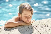 Desde arriba de un lindo niño reflexivo apoyado en el borde de la piscina mientras descansa después de nadar en un día soleado - foto de stock