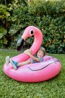 Corpo pieno di allegro bambino in abiti casual seduto su fenicottero rosa gonfiabile mentre si diverte sul prato erboso nel parco — Foto stock