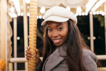 Side view of African American female on merry go round smiling at camera while entertaining in amusement park in evening — Stock Photo