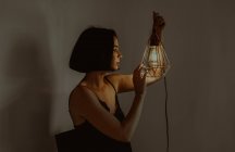 Side view of delicate female touching illuminated metal lantern while sitting in room at home — Stock Photo