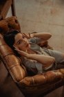 From above of peaceful female sitting in shabby leather chair and touching head while relaxing at home and looking at camera — Stock Photo