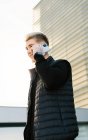 Low angle side view of happy young male with stylish haircut in casual clothes smiling while having phone conversation on city street near modern building at sunset — Stock Photo