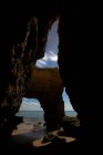 Through cave scenery view of rough rocky formations on sandy Pinhao Beach against sea under cloudy sky in Algarve Portugal — Stock Photo