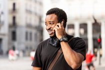 Joven contenido afroamericano hombre en reloj de pulsera hablando por teléfono celular mientras mira hacia otro lado en la ciudad - foto de stock