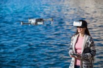 Excited female wearing modern VR goggles operating drone with remote controller and experiencing virtual reality while standing against blurred sea — Stock Photo