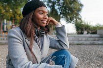 Carefree African American female in outerwear sitting on street and leaning on hand while looking away in contemplation — Stock Photo