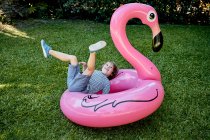 Все тело веселого маленького мальчика в повседневной одежде лежит на надувном розовом фламинго, веселясь на травянистой лужайке в парке — стоковое фото