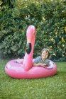 Corps complet de gai petit garçon en vêtements décontractés couché sur flamant rose gonflable tout en s'amusant sur pelouse herbeuse dans le parc — Photo de stock