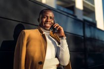 Concentrato giovane afroamericano maschio in elegante dolcevita e cappotto parlando sul telefono cellulare e guardando lontano pensieroso mentre in piedi sulla strada della città — Foto stock
