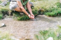 Полное тело обрезали неузнаваемая женщина-турист в повседневной одежде мыть руки в ручье, сидя на травянистом лугу в каменистой местности — стоковое фото