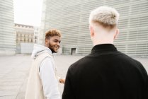 Vista lateral de jovens amigos homens multirraciais felizes em roupas da moda em pé na rua da cidade perto do edifício moderno — Fotografia de Stock