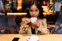 Langhaarige brünette asiatische Frau, die einen Kaffee in einem Coffeeshop trinkt, während sie ein Handy sucht — Stockfoto