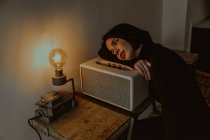 Високий кут схилення мрійливої жінки на радіо встановлений у ретро-кімнаті і дивиться на лампочку з підсвічуванням — стокове фото