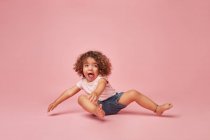 Cute cheerful toddler girl with curly hair in casual clothes having fun making faces while sitting on the floor looking away on pink background — Stock Photo