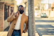 Неузнаваемый вдумчивый молодой чернокожий мужчина в модном наряде и маске для лица, опирающийся на дерево на городской улице и отворачивающийся, разговаривая по телефону — стоковое фото